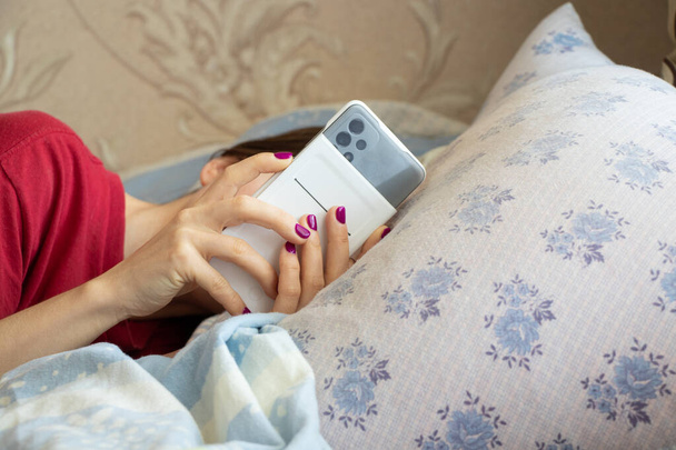 Μια νεαρή κοπέλα με ένα τηλέφωνο στα χέρια της βρίσκεται στο κρεβάτι, μιλώντας στο τηλέφωνο στο κρεβάτι, χαλαρώνοντας στο σπίτι - Φωτογραφία, εικόνα