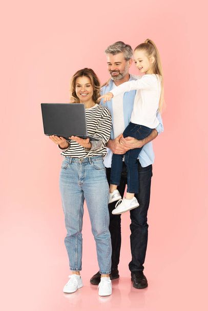Familia feliz usando el ordenador portátil que navega Internet juntos, hija pequeña que señala el dedo en la computadora que recomienda el Web site que posa sobre fondo rosado del estudio. Tiro vertical - Foto, Imagen