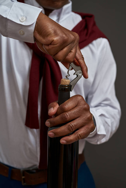 Cropped of african american winemaker or sommelier opening wine bottle with corkscrew. Successful male entrepreneur or businessman. Viticulture and winemaking. Isolated on grey background in studio - Foto, Bild