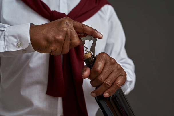 Partial of black winemaker or sommelier opening wine bottle with corkscrew. Successful male entrepreneur or businessman. Viticulture and winemaking. Isolated on grey background. Studio shoot - Fotó, kép