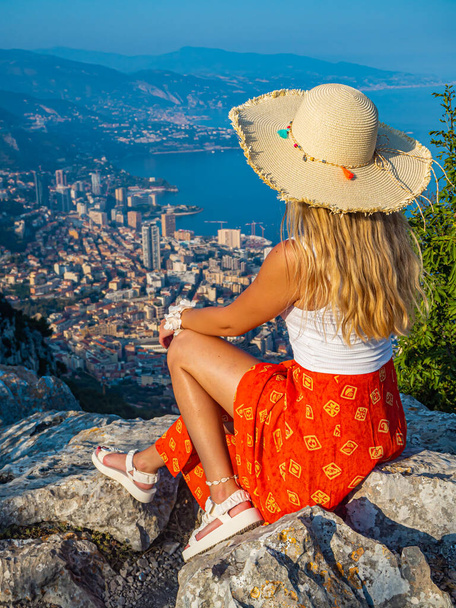 Giovane ragazza bionda con un cappello seduta sul bordo di una montagna con una splendida vista panoramica di Monaco durante il sole al tramonto, Monte Carlo, Costa Azzurra - Foto, immagini