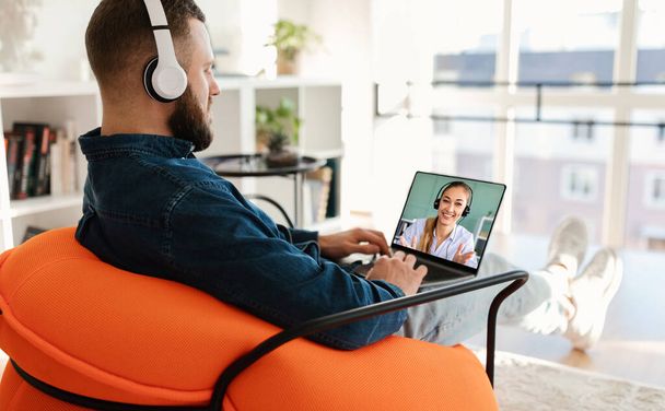 Millennial Guy usando laptop, vestindo fones de ouvido sem fio, tendo videoconferência com parceira de negócios feminina, trabalhando on-line sentado na cadeira dentro de casa. Conceito de trabalho freelance e à distância, espaço de cópia - Foto, Imagem