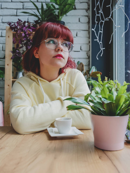 Stylish redhead teenager girl with cup of coffee sit at table in cafe with plants around - Zdjęcie, obraz