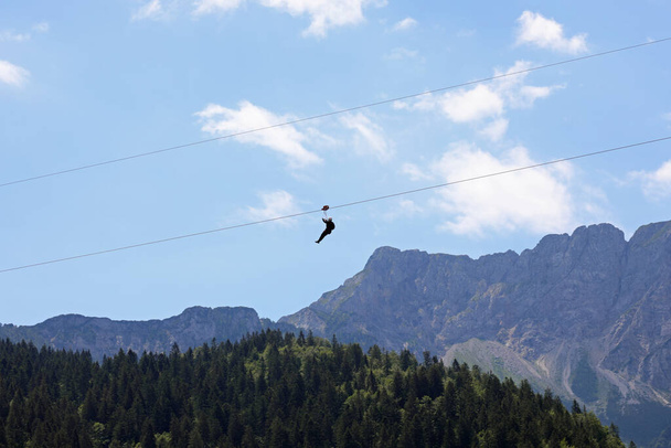 Бесстрашный человек висит на стальном кабеле, который соединяет противоположные склоны горы и силу тяжести, которая тянется вниз и горы на заднем плане - Фото, изображение