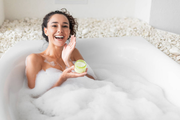 Lovely young woman lying in warm bubble bath, applying cream or face mask, copy space. Attractive millennial female relaxing in bathtub, taking care of her skin at luxury hotel - Photo, Image