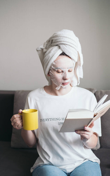 mujer joven está sentada en el sofá en una toalla y una máscara cosmética y leyendo un libro - Foto, imagen