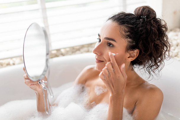 Veselá tisíciletá žena používající hydratační krém na obličej ve vaně, dívající se do zrcadla doma. Šťastná mladá žena nanášení vyživující krém na obličej při relaxaci v bublinkové koupeli uvnitř - Fotografie, Obrázek