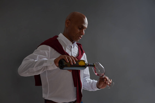 Concentrated male adult black winemaker or sommelier pouring red wine in glass from bottle. Wine tasting. Viticulture and winemaking. Isolated on grey background. Studio shoot. Copy space - Photo, image