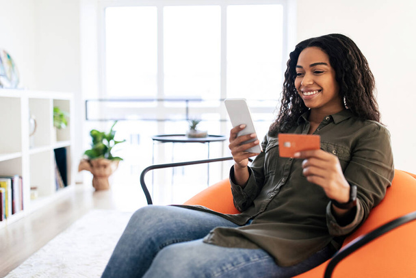 E-Commerce-Konzept. Lächelnde schwarze Dame, die ihre EC-Kreditkarte mit dem Handy hält und auf den Bildschirm schaut, Kunde, der Finanztransaktionen tätigt, sitzt auf Sitzsack-Sessel, verschwommener Hintergrund, freier Kopierraum - Foto, Bild