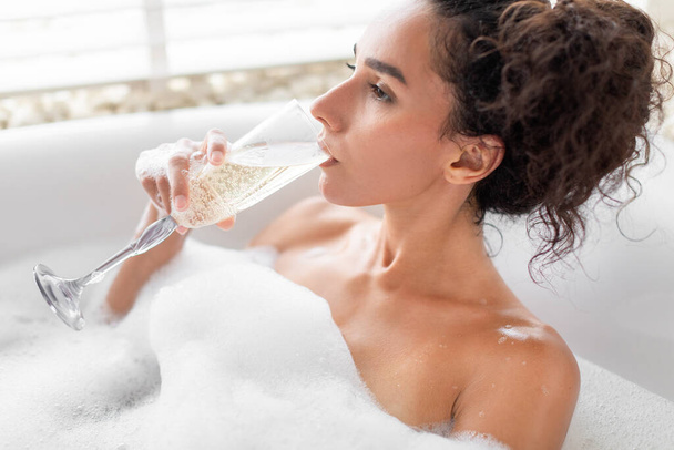 Dreamy millennial lady drinking champagne while relaxing in bubble bath at luxury hotel. Closeup portrait of thoughtful young woman enjoying alcoholic beverage in bathtub at home - Photo, Image