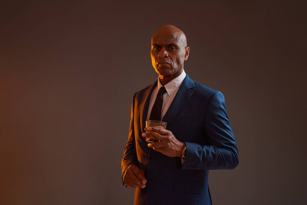 Confident black entrepreneur hold cigar and glass with whiskey or brandy. Adult man wear formal wear. Modern successful male lifestyle. Isolated on dark red with light background in studio. Copy space - Foto, afbeelding
