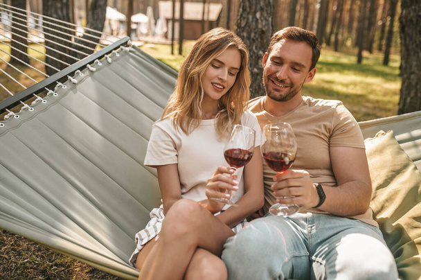 Smiling pleased beautiful blonde woman and her happy boyfriend sitting in the hammock and clinking glasses - Valokuva, kuva