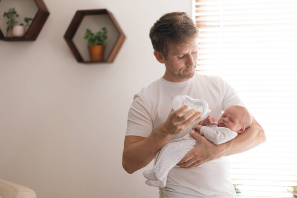 A father holding and feeding his newborn son with milk bottle at home. - Photo, Image