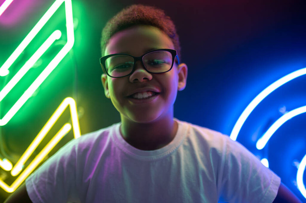 Portrait of a smiling cheerful African American teenage boy in fashionable eyeglasses in a nightclub - 写真・画像