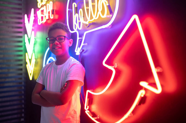 Waist-up portrait of a happy teenage boy in trendy spectacles posing for the camera in a nightclub - Fotó, kép