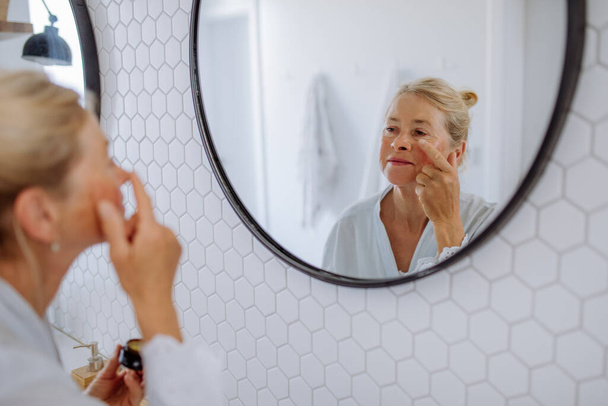 Красива старша жінка в халаті, що застосовує натуральний крем для обличчя у ванній кімнаті, догляді за шкірою та ранковій рутині концепції
. - Фото, зображення