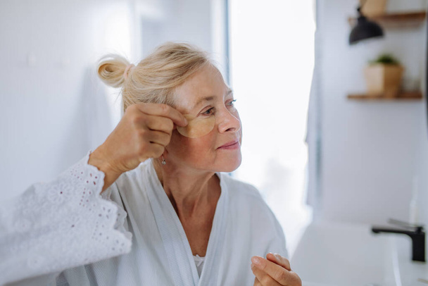 A beautiful senior woman in bathrobe, applying eye patches for puffiness while looking in the mirror - Foto, Imagen