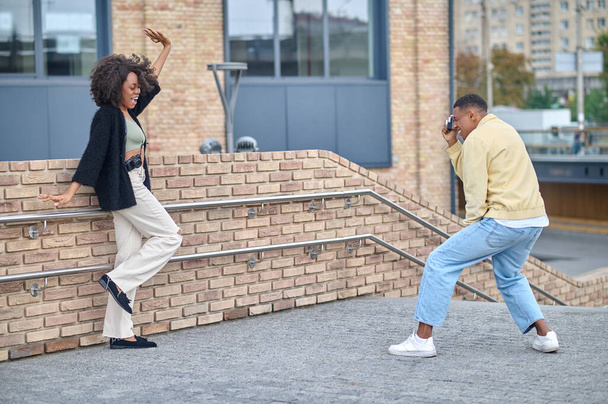 Inspiration. African american guy standing sideways to camera photographing cheerful girl with open mouth in strike pose on city street - Foto, afbeelding