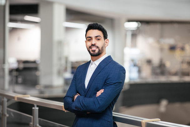 Smiling confident calm attractive young middle eastern businessman with beard in suit with crossed arms looks at camera in office interior. Successful business, ceo manager and leadership at factory - Zdjęcie, obraz