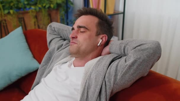 Happy young man wearing wireless earphones listening favorite energetic disco music song, relaxing, sleeping, resting, having a break, lying on couch at home. Weekends entertaining activities indoors - Footage, Video