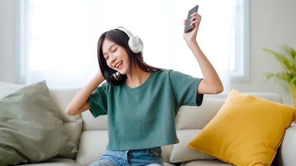 Fiatal ázsiai nő zenét hallgat a kanapén otthon a nappaliban. Boldog ázsiai nő használ mobil okostelefon, visel headset és ül a kanapén - Fotó, kép