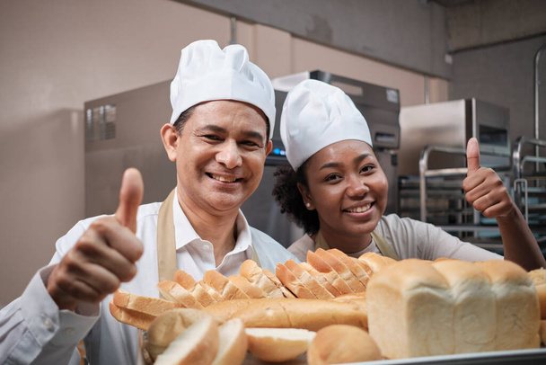 Portret van professionele chef-koks in wit uniform kijkend naar camera met vrolijke glimlach en duim omhoog met dienblad brood in de keuken. Vriend en partner van bakkerijproducten en verse dagelijkse bakkerij beroep. - Foto, afbeelding