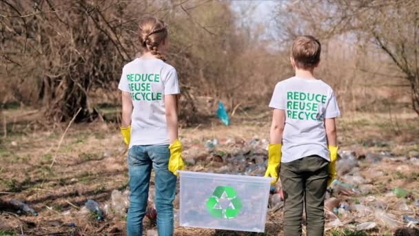 Chłopiec i dziewczyna w gumowych rękawiczkach trzymający pojemnik na plastikowym śmietniku na zanieczyszczonej polanie, znaki recyklingu na koszulkach. Zwolniony ruch - Materiał filmowy, wideo