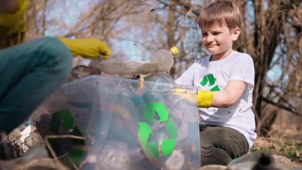 Fiú és lány műanyag szemetet gyűjt egy konténerben egy szennyezett tisztáson, újrahasznosító táblákkal a pólókon. Lassú mozgás. - Felvétel, videó