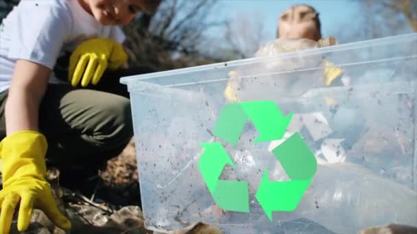 Jongen en meisje verzamelen plastic afval in een container in een vervuilde open plek, recycling tekens op de T-shirts. Langzame beweging - Video