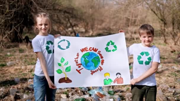 Jongen en meisje houden poster ter verdediging van de ecologie bij plastic vuilnisophaling in een vervuilde open plek, recycling tekens op de T-shirts. Langzame beweging - Video