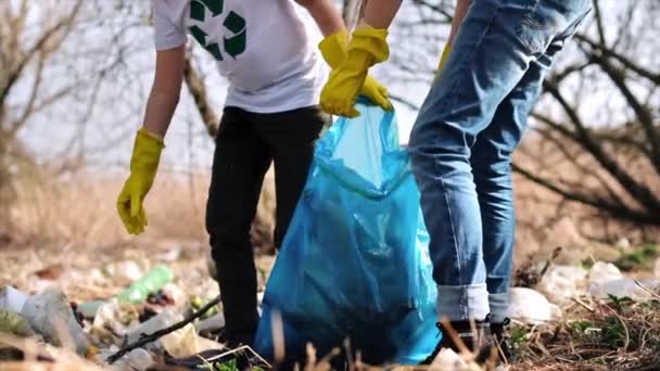Jongen en meisje verzamelen plastic afval in een zak in een vervuilde open plek, recycling tekens op de T-shirts. Langzame beweging - Video