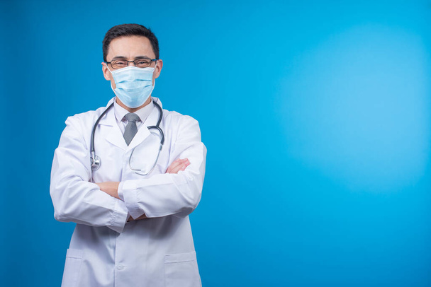 Содержательный врач в защитной маске скрещивает руки и смотрит в камеру со стетоскопом на шее на синем фоне - Фото, изображение