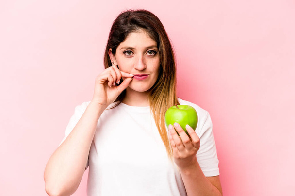 Mujer caucásica joven sosteniendo una manzana aislada sobre fondo rosa con los dedos en los labios manteniendo un secreto. - Foto, Imagen