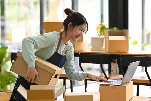 Lächelnde junge asiatische Online-Verkäuferin, die ihre Bestellung am Laptop überprüft. E-Commerce, Online-Geschäft, Online-Vertriebskonzept. - Foto, Bild