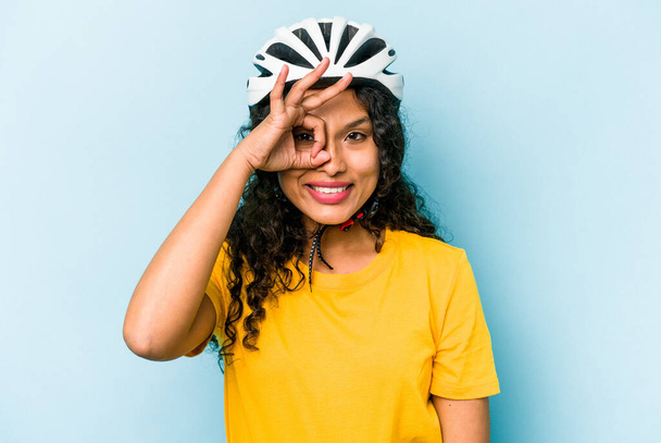 Młoda Latynoska kobieta nosząca rower w kasku odizolowana na niebieskim tle podekscytowana trzymając ok gest na oku. - Zdjęcie, obraz