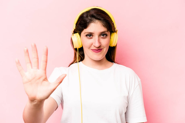 Young caucasian woman listening to music isolated on pink background smiling cheerful showing number five with fingers. - Fotografie, Obrázek