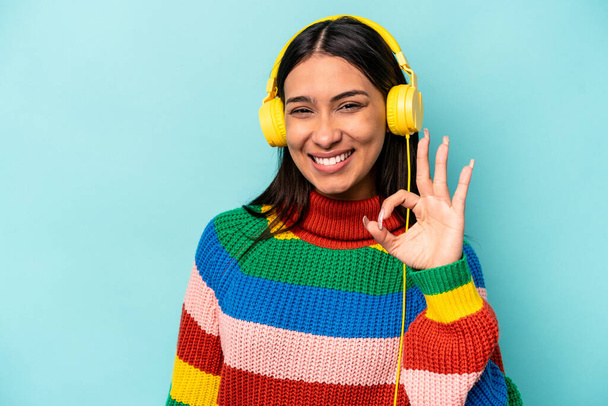 Jonge Spaanse vrouw luisteren naar muziek geïsoleerd op blauwe achtergrond vrolijk en zelfverzekerd tonen ok gebaar. - Foto, afbeelding