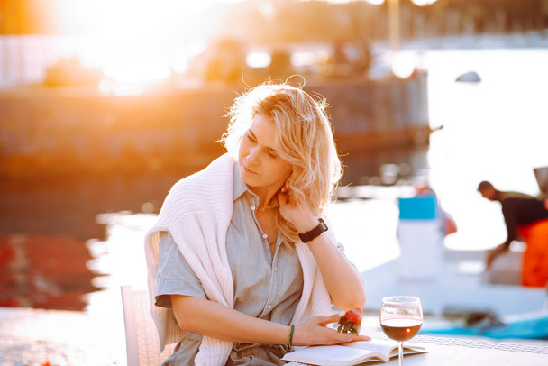 Portrait of young beautiful woman with long wavy fair hair, wearing blue shirt, white sweater, sitting at table with glass of red wine and book, holding strawberry in hand on embankment at sunset. - Foto, Imagen