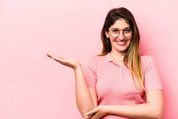 Νεαρή καυκάσια γυναίκα απομονωμένη σε ροζ φόντο που δείχνει ένα χώρο αντιγραφής σε μια παλάμη και κρατώντας ένα άλλο χέρι στη μέση. - Φωτογραφία, εικόνα