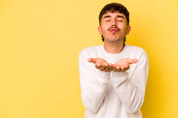 Young caucasian man isolated on yellow background folding lips and holding palms to send air kiss. - Foto, immagini