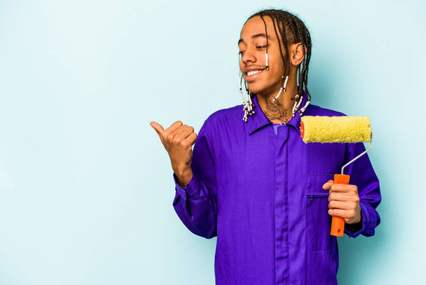 Young African American painter man isolated on blue background points with thumb finger away, laughing and carefree. - Photo, Image