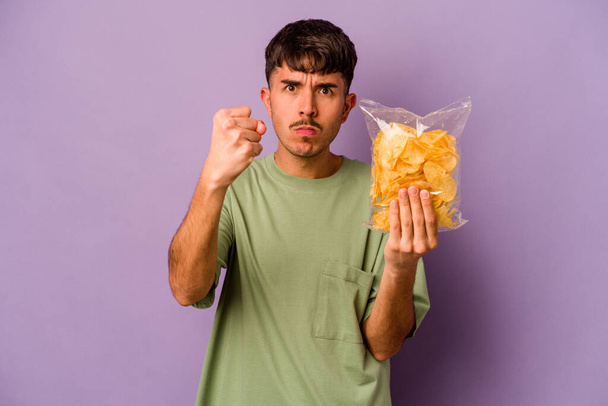 Joven hispano sosteniendo patatas fritas aisladas sobre fondo púrpura mostrando puño a cámara, agresiva expresión facial. - Foto, imagen