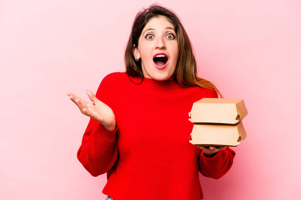Young caucasian woman holding burgers isolated on pink background surprised and shocked. - 写真・画像