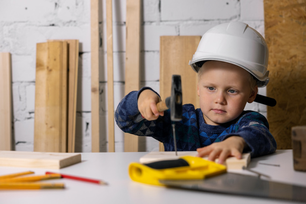 Маленький мальчик в шлеме учится забивать гвоздь в деревянную доску в мастерской плотников - Фото, изображение