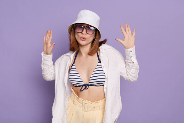 Horizontal shot of playful cheerful young adult woman wearing swimsuit, shirt, shorts and panama, standing with raised arms and pout lips, posing isolated over purple background. - Fotoğraf, Görsel