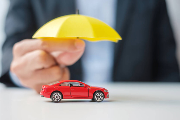 Empresario mano sosteniendo paraguas y cubierta de juguete de coche rojo en la mesa. Seguro de automóvil, garantía, reparación, concepto financiero, bancario y monetario - Foto, imagen