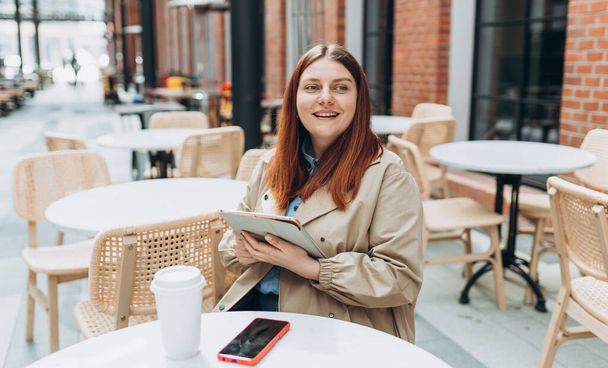 Νεαρή γυναίκα απολαμβάνει έναν καφέ, κάθεται στη βεράντα του καφέ στο σύγχρονο δρόμο της πόλης. Άτομο που κάθεται στο τραπέζι και χρησιμοποιεί δισκίο σε εξωτερικούς χώρους. Traveler, Online εκπαίδευση, έννοια εργασίας - Φωτογραφία, εικόνα
