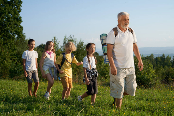 Side view of old man leading group of kids behind him in woods. Male with gray hair and beard, wearing rucksack showing route to four children. Concept of active rest and hiking. - Φωτογραφία, εικόνα