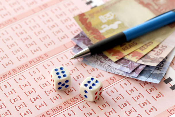 Kleine Würfel mit brasilianischen Geldscheinen und Stift auf dem Rohling des Lotteriespiels. Konzept von Glück und Glücksspiel in Brasilien - Foto, Bild