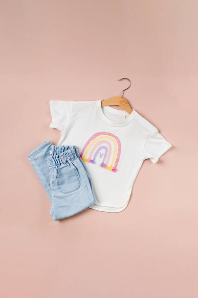 Childrens t-shirt with a rainbow and folded jeans on a pink background. Childrens fashion image. Flat lay. - Foto, Bild